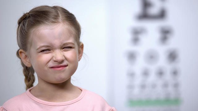 Small girl frowning, trying to see letter on vision testing table blurred vision 